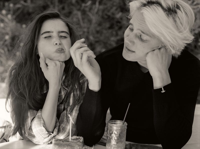 A young woman with long hair eating a piece of cake while a person with short blonde hair watches her in a sepia-toned shot.