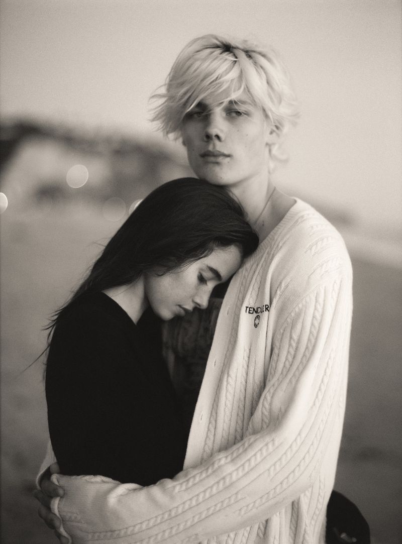 Monochrome portrait of a blonde young man in an off-white cable-knit cardigan embracing a dark-haired young woman outdoors.