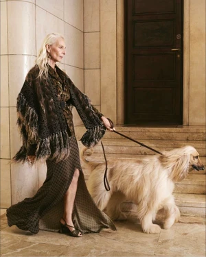 Model Elisabetta Dessy in a fringed brown patterned outfit walking an Afghan hound dog beside stone stairs.