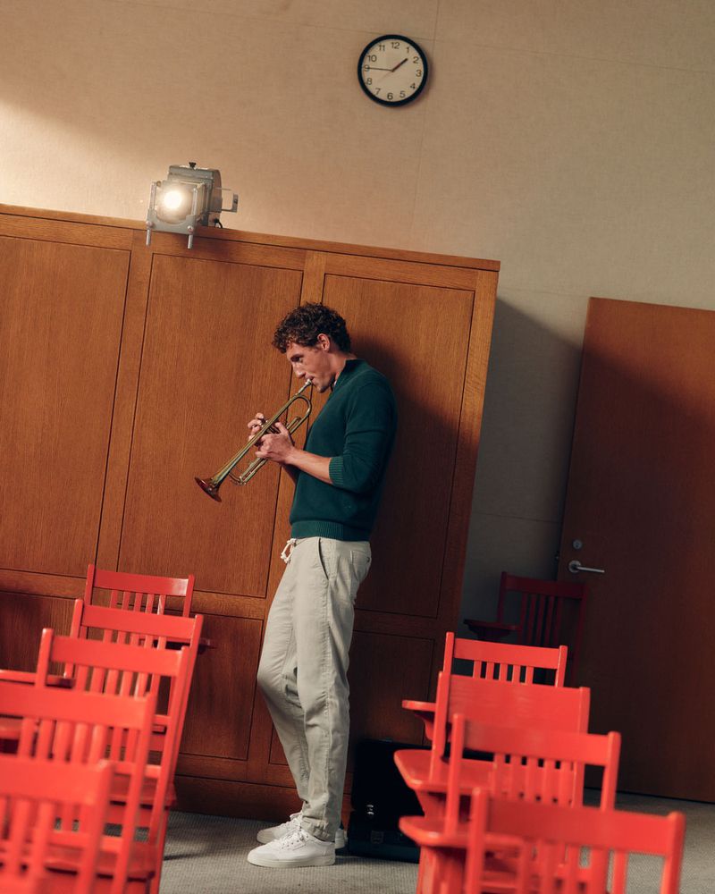 Man with curly brown hair playing a trumpet while standing in a room with orange chairs and wooden walls