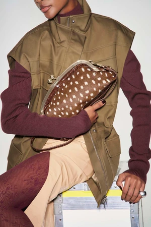 Model holding a brown spotted clutch handbag while wearing an olive khaki utility vest and burgundy knit sweater.