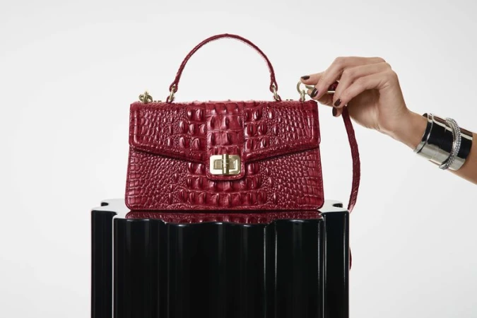 Red croc-embossed leather handbag resting on a black fluted pedestal, held by a woman's hand wearing silver cuffs.