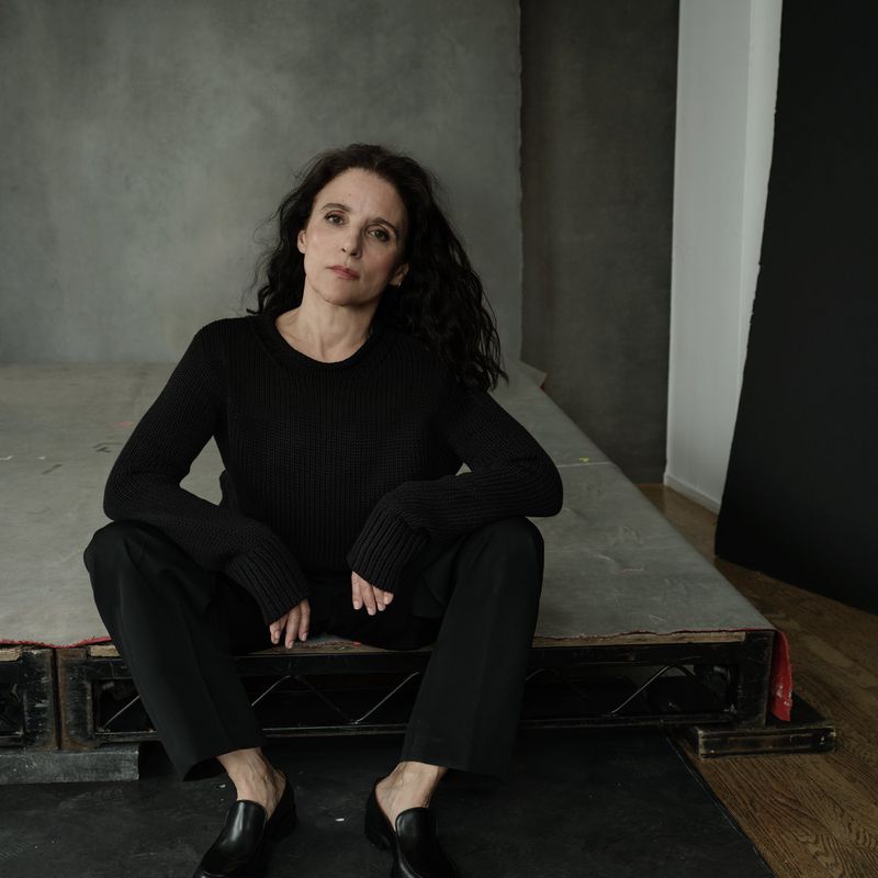 Julia Louis-Dreyfus seated on a wooden platform wearing a black long-sleeved sweater, black trousers, and black loafers.