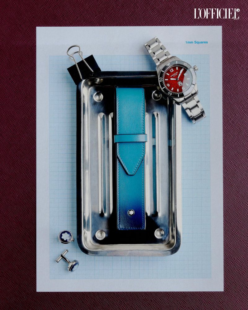 Elevated shot of a wristwatch, turquoise leather strap, cufflinks, and binder clip on a metal tray, set on graph paper against a red background.