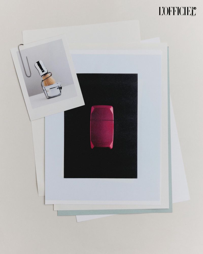 Still life of photographs and paper, featuring a pink cosmetic case and a shaving brush attached with a silver paper clip.