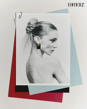 Studio shot of a bare-shouldered woman wearing large square earrings, styled in a bun, in front of overlapping blue, red and black paper.