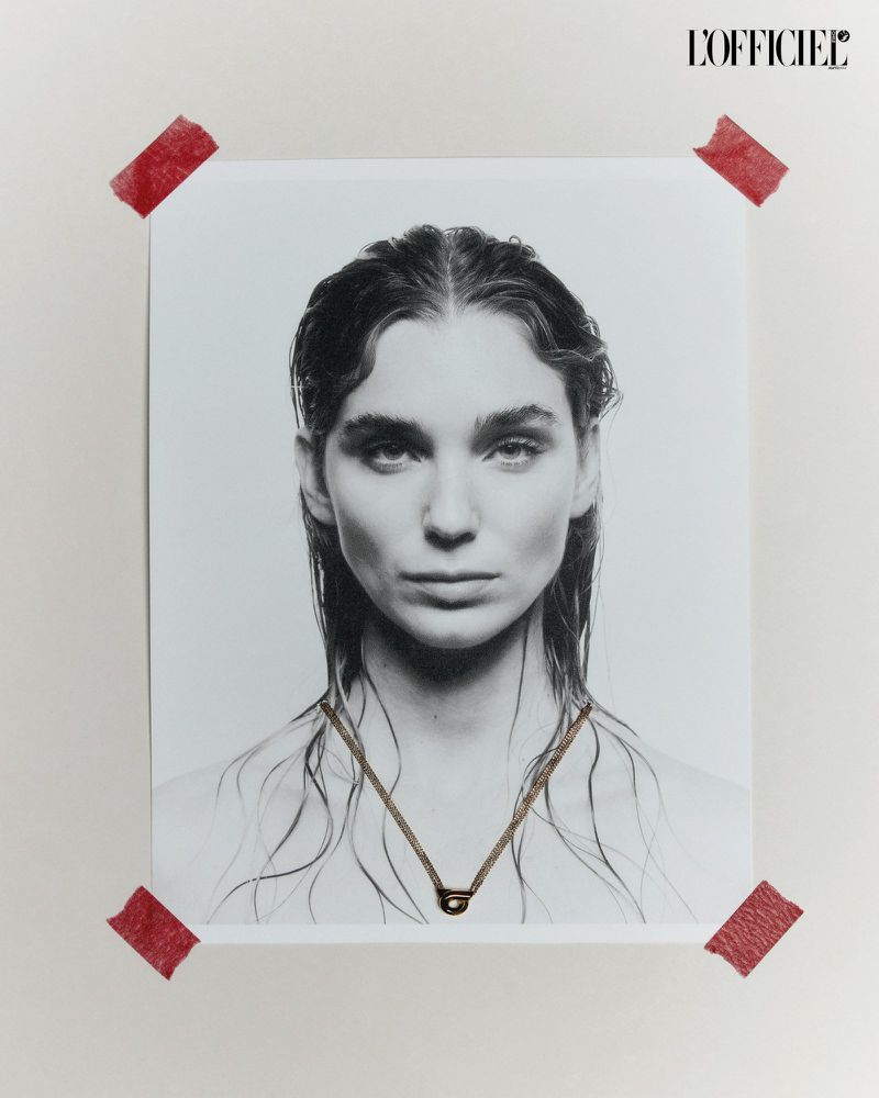 Black and white close-up portrait of a woman with wet hair and a gold necklace attached to a white paper taped to a white wall.