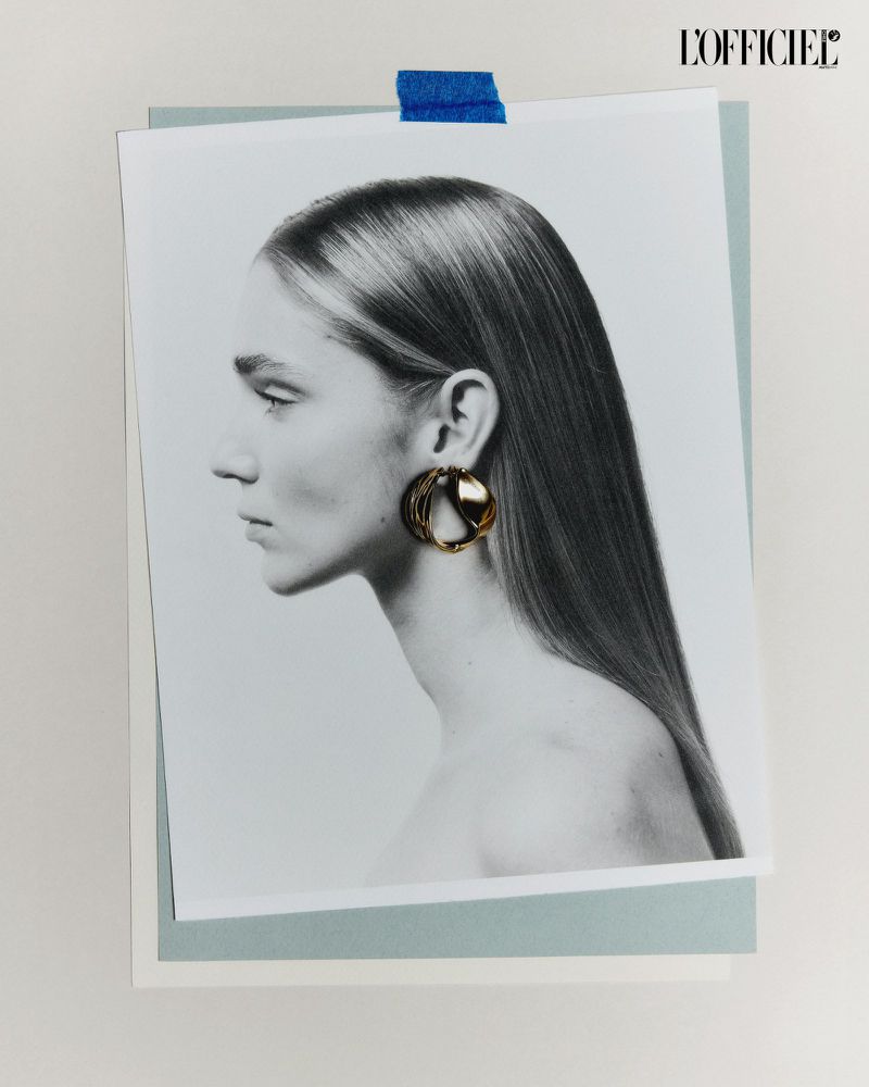 Monochrome portrait of a blonde woman in profile wearing gold hoop earrings, taped to a blue and white layered paper background.