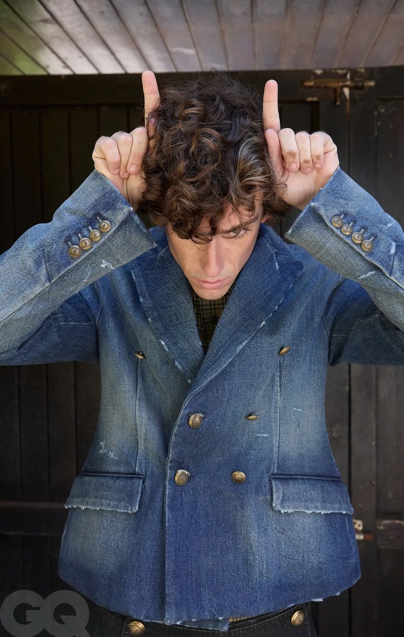 A young man with curly hair wearing a blue denim double-breasted jacket making a hand gesture above his head against a dark wooden door.