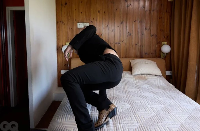 Guitarricadelafuente crouching on a white quilted bed wearing a black suit and snakeskin boots in a wood-paneled room.