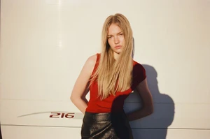Blonde woman in a red top and black textured leather skirt leaning against a white trailer panel under direct sunlight.