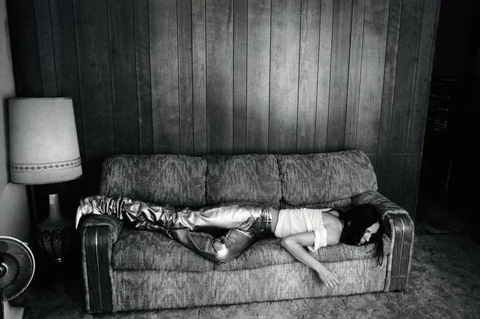 Model Waleska Gorczevski wearing metallic silver pants lying prone across a patterned vintage sofa against a wood paneled wall.
