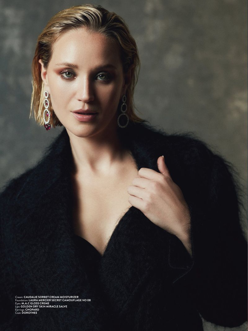 Close-up portrait of a woman with wet-look blonde hair wearing long silver earrings and a black textured coat against a grey backdrop.