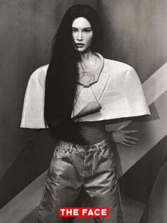 Model Sui He wearing a sculptural wide-shoulder cropped jacket and denim jeans against a geometric studio background.