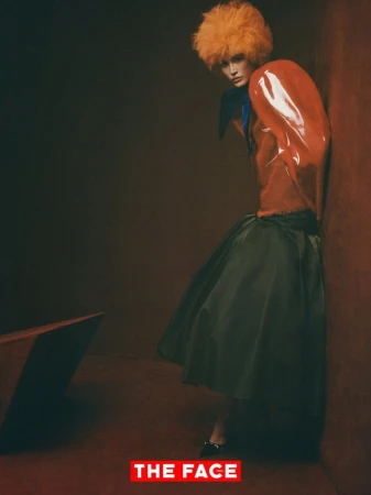 Model Sui He in an orange wig, glossy vinyl top, and green flared skirt leaning against a studio corner wall.