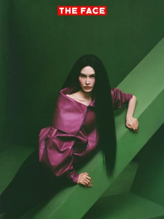 Model Sui He wearing an oversized magenta leather jacket with long black hair, leaning across a green geometric set piece.