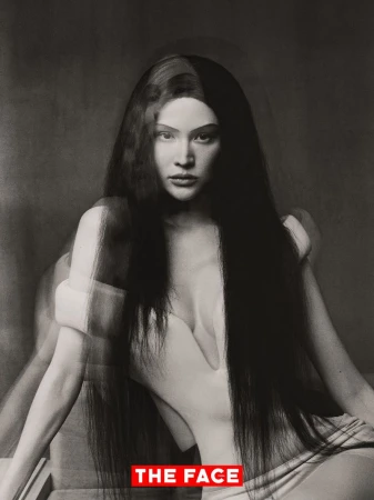 Model Sui He seated in a sculptural white off-shoulder dress with a motion blur photographic effect against a studio backdrop.