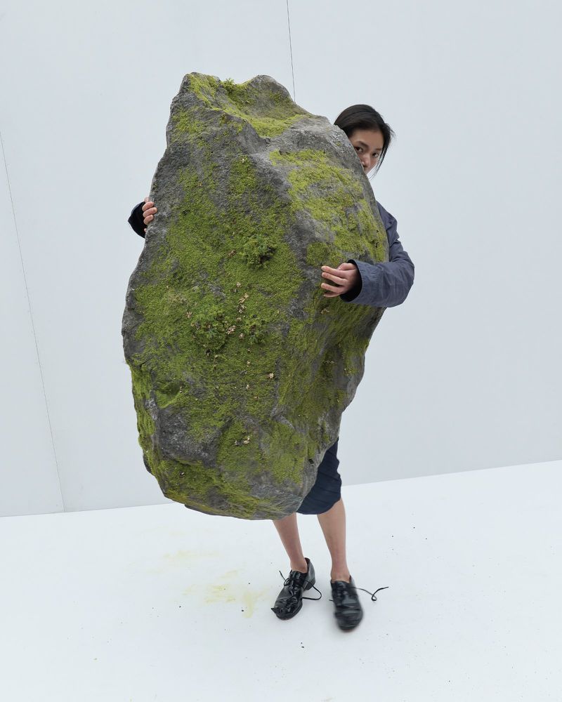 A model standing in a white studio holding a massive moss-covered grey boulder in front of her body.