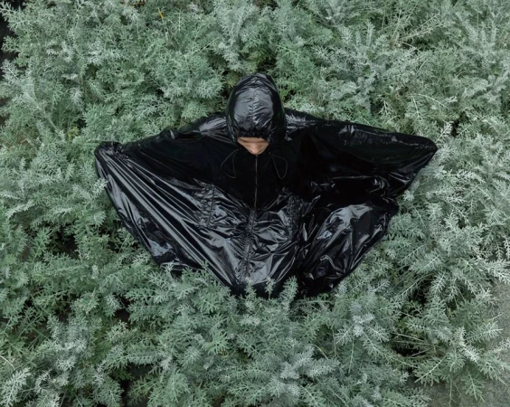 High-angle view of a model wearing a glossy black hooded cape seated in dense silvery green foliage.