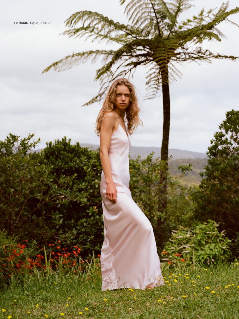 Model Sabine Glud wears a long blush pink satin slip dress while standing outdoors near a tree fern on a grassy slope.