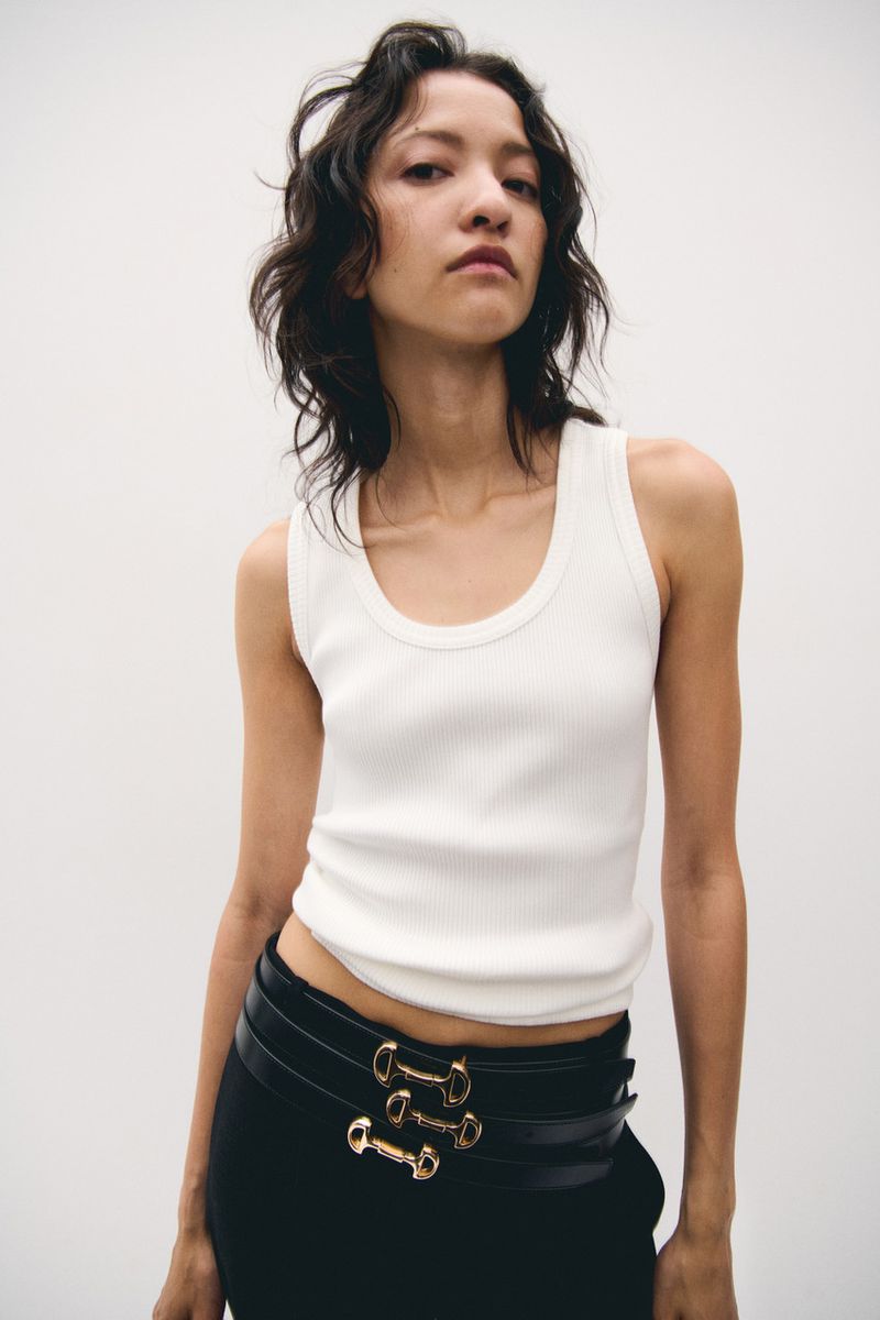 Fashion portrait of a young adult female with wavy dark hair wearing a white tank top and black pants with a gold belt.