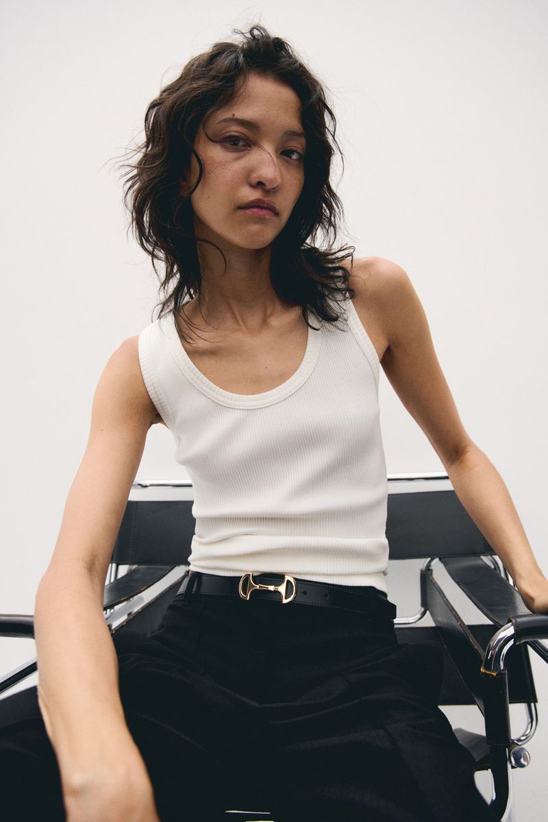 Close-up of a woman in a white tank top sitting on a black chair, posing indoors under bright lighting with a neutral background.