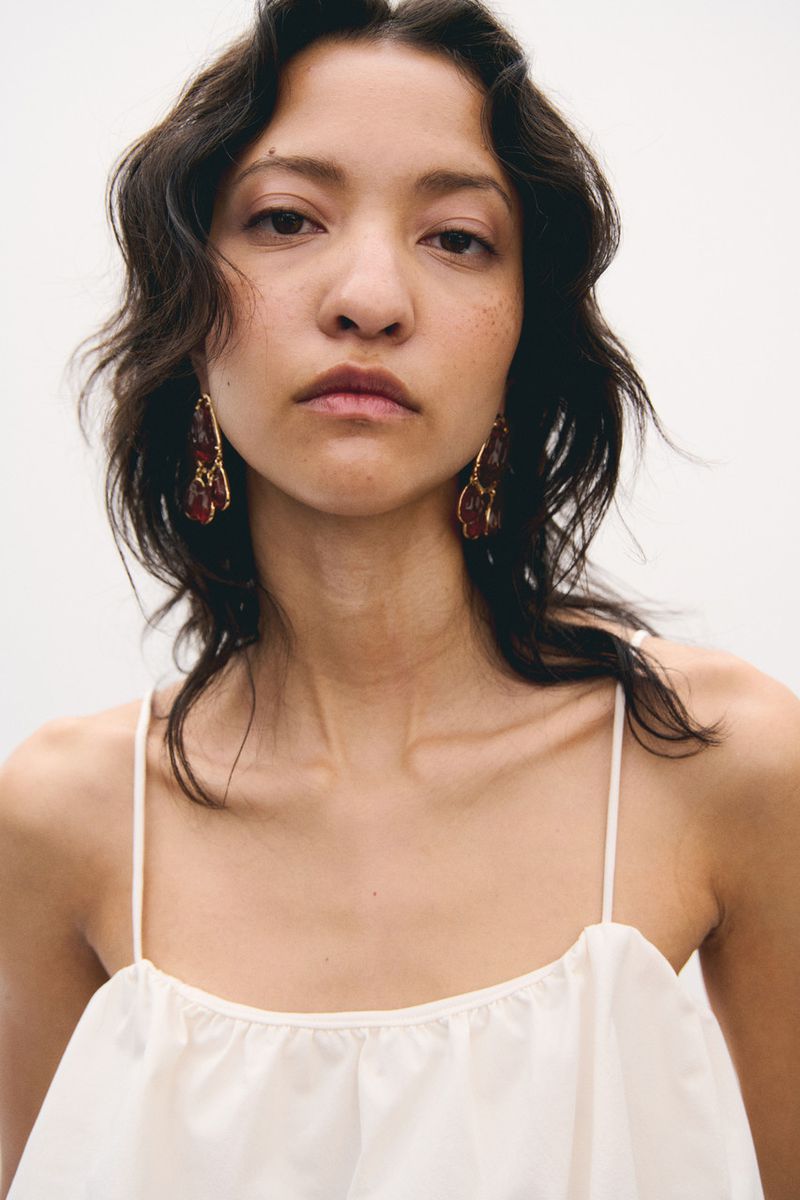 Close-up portrait of a young woman with dark wavy hair wearing a light-colored top and chandelier earrings against a white backdrop.