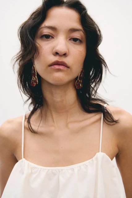 Close-up portrait of a young woman with dark wavy hair wearing a light-colored top and chandelier earrings against a white backdrop.