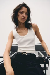 Model América González in a white tank top and black pants seated on a chair against a white background for H&M.