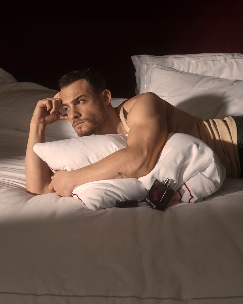 Man lying on a white bed holding a small perfume bottle, wearing a beige tank top, looking pensively toward the side.