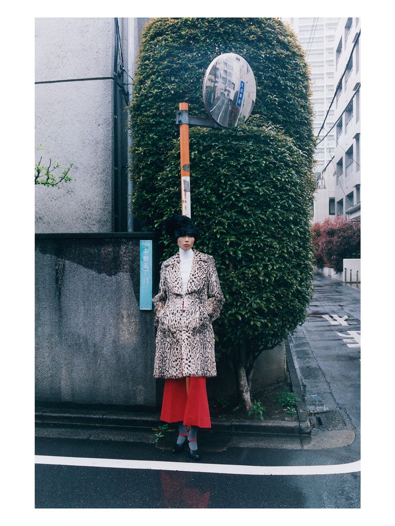 A person standing on an urban street wearing a leopard print coat, white turtleneck, and long red skirt near a tall hedge.