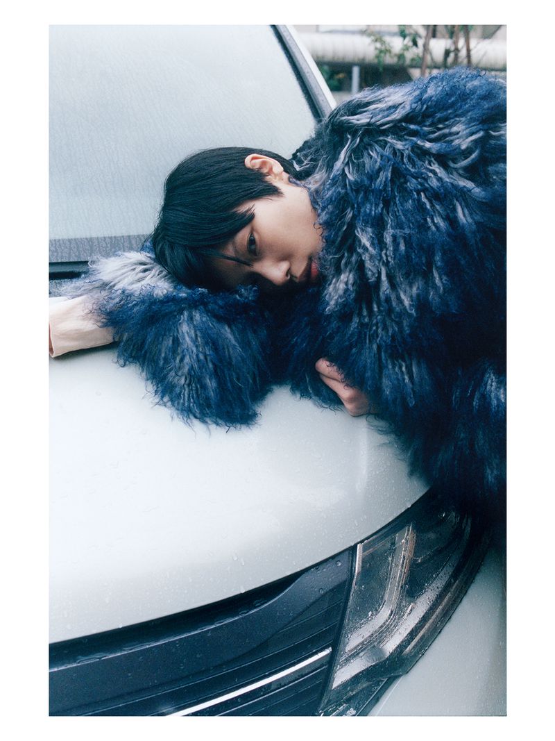 Model Kayako Higuchi lying across the hood of a car wearing a shaggy blue faux fur coat.