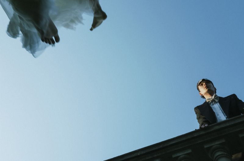 A man in a tuxedo stands on a balcony looking up at the bare feet and flowing white fabric of a figure floating in the sky above.