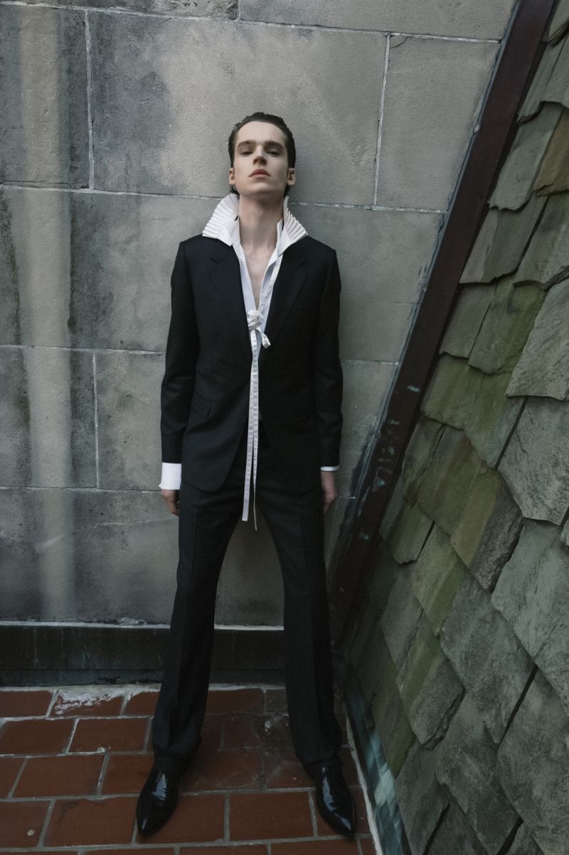 Male model posing in a black suit with an exaggerated white pleated collar shirt against an outdoor stone wall.