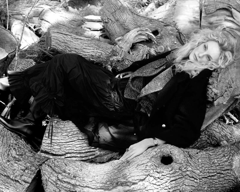 Blonde woman wearing a dark coat and ruffled trousers lying on a pile of fallen logs in a black and white editorial style photograph.