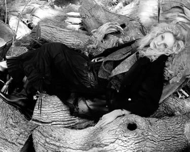 Model lying across a pile of fallen tree logs wearing a dark coat, patterned vest, and ruffled trousers in black and white.