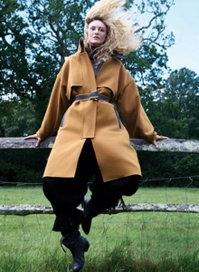 Model Caroline Trentini wearing a belted mustard yellow coat perched on a wooden fence in a grassy field with windblown hair.