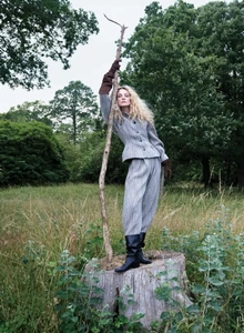 Model posing on a tree stump in a grey tweed suit and black boots while holding a tall wooden branch in a grassy woodland.
