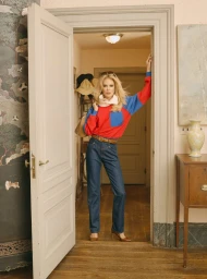 Blonde woman in a red and blue colorblock top and high-waisted denim jeans posing in an open interior doorway.