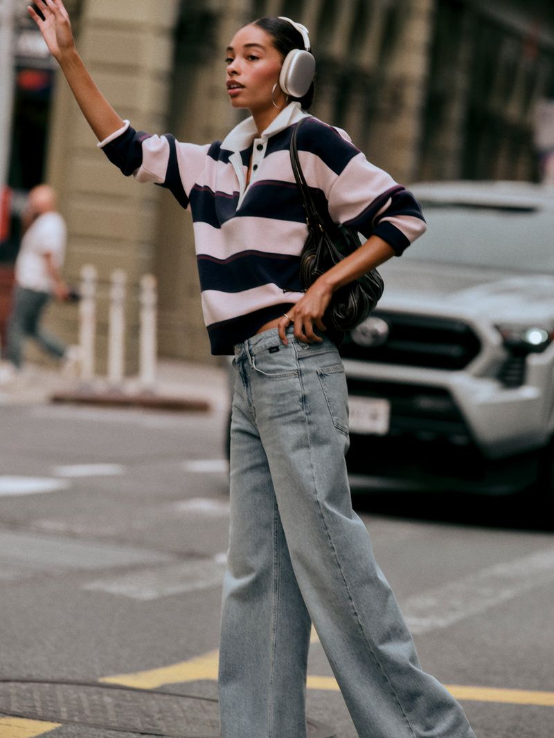 Model wearing wide-leg light wash denim jeans and a striped rugby sweater with over-ear headphones standing on a city street.