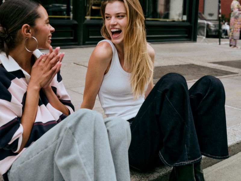 Two women seated on a city sidewalk laughing together, one in a white tank top and dark jeans, the other in a striped rugby shirt.
