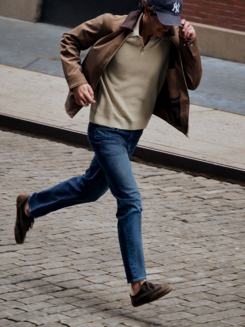 Man in blue denim jeans and brown jacket running across a cobblestone street wearing a navy baseball cap.