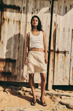Model Alba Martin in a white Ibiza tank top and cream miniskirt standing against a weathered wooden wall.