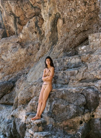 Model wearing a striped bikini standing barefoot on rugged stone steps against a sheer coastal rock cliff.