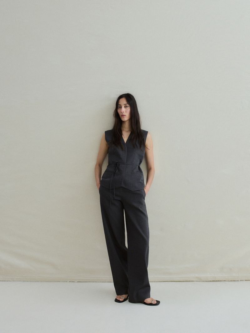 A woman in a dark grey sleeveless jumpsuit stands with her hands in her pockets against a light background, creating a minimalist fashion shot.