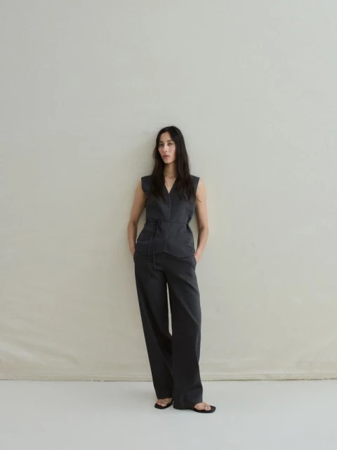A woman in a dark grey sleeveless jumpsuit stands with her hands in her pockets against a light background, creating a minimalist fashion shot.