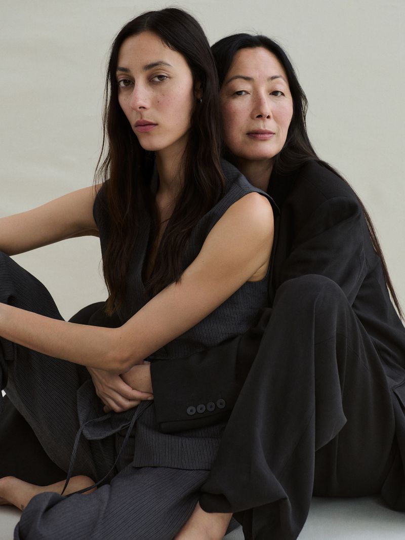 Two Asian women in dark suits pose together indoors. The younger woman wears a vest; the other wears a suit jacket and wide-leg pants.