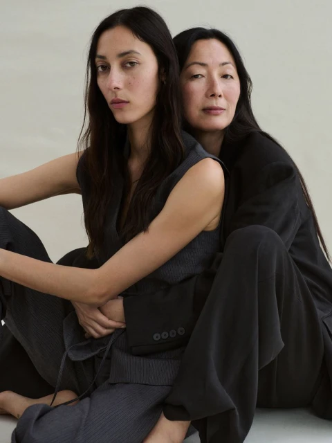 Two Asian women in dark suits pose together indoors. The younger woman wears a vest; the other wears a suit jacket and wide-leg pants.