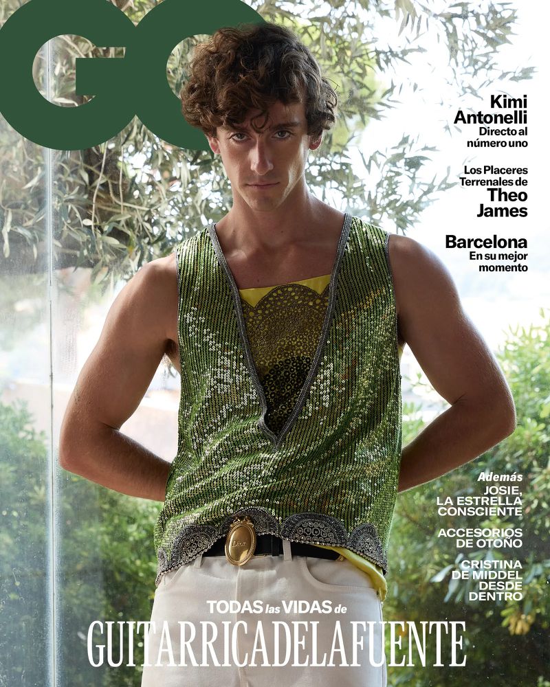 Young man with curly brown hair wears a green sequined sleeveless top and white trousers on a magazine cover against foliage.