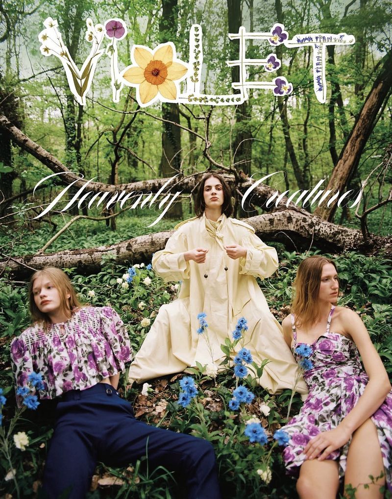 Three models in floral and pastel clothing posed among wildflowers and fallen trees in a lush, green forest.